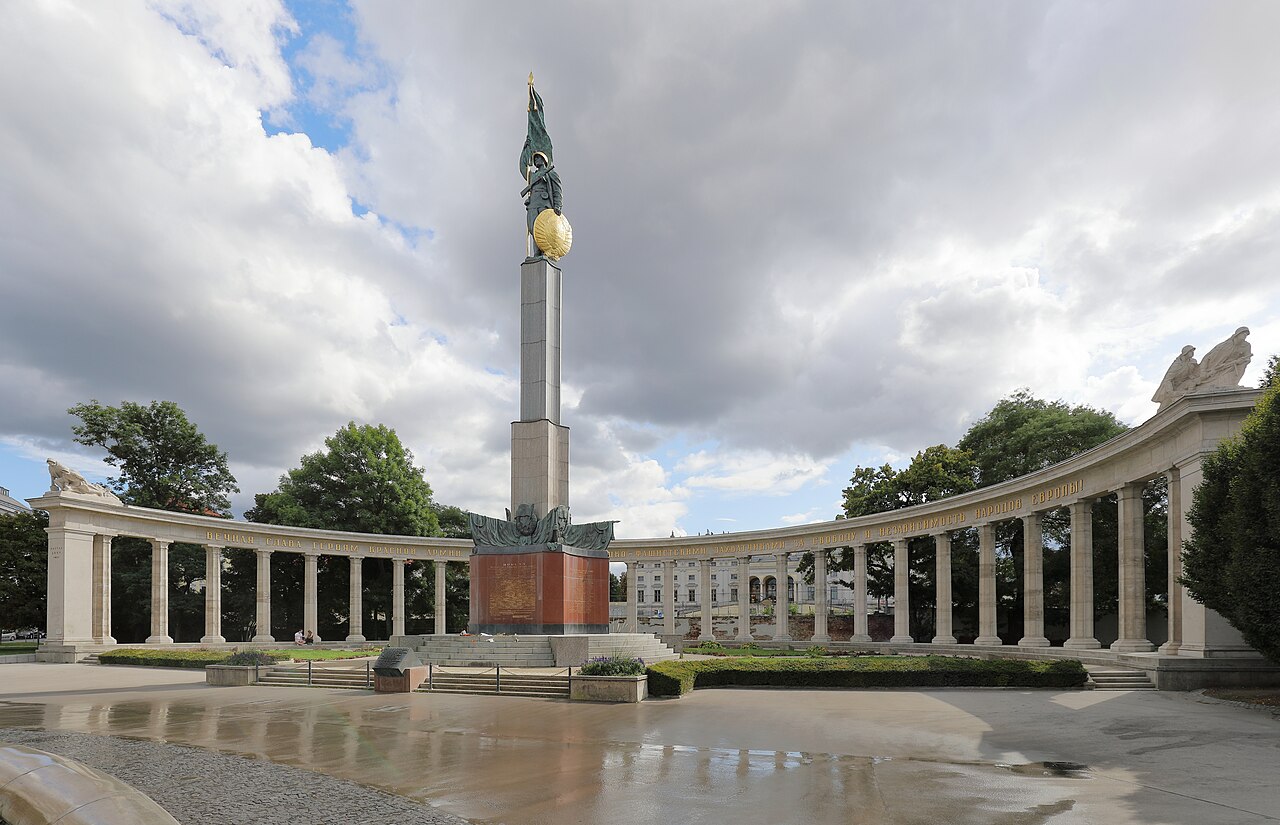 A tall column topped by a soldier with a flag and a gilded shield, in front of a curved colonnade.