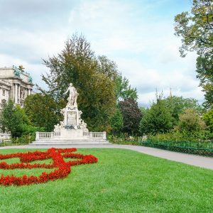 A white marble statue on a tall base above a lawn with a treble clef planted in red flowers.